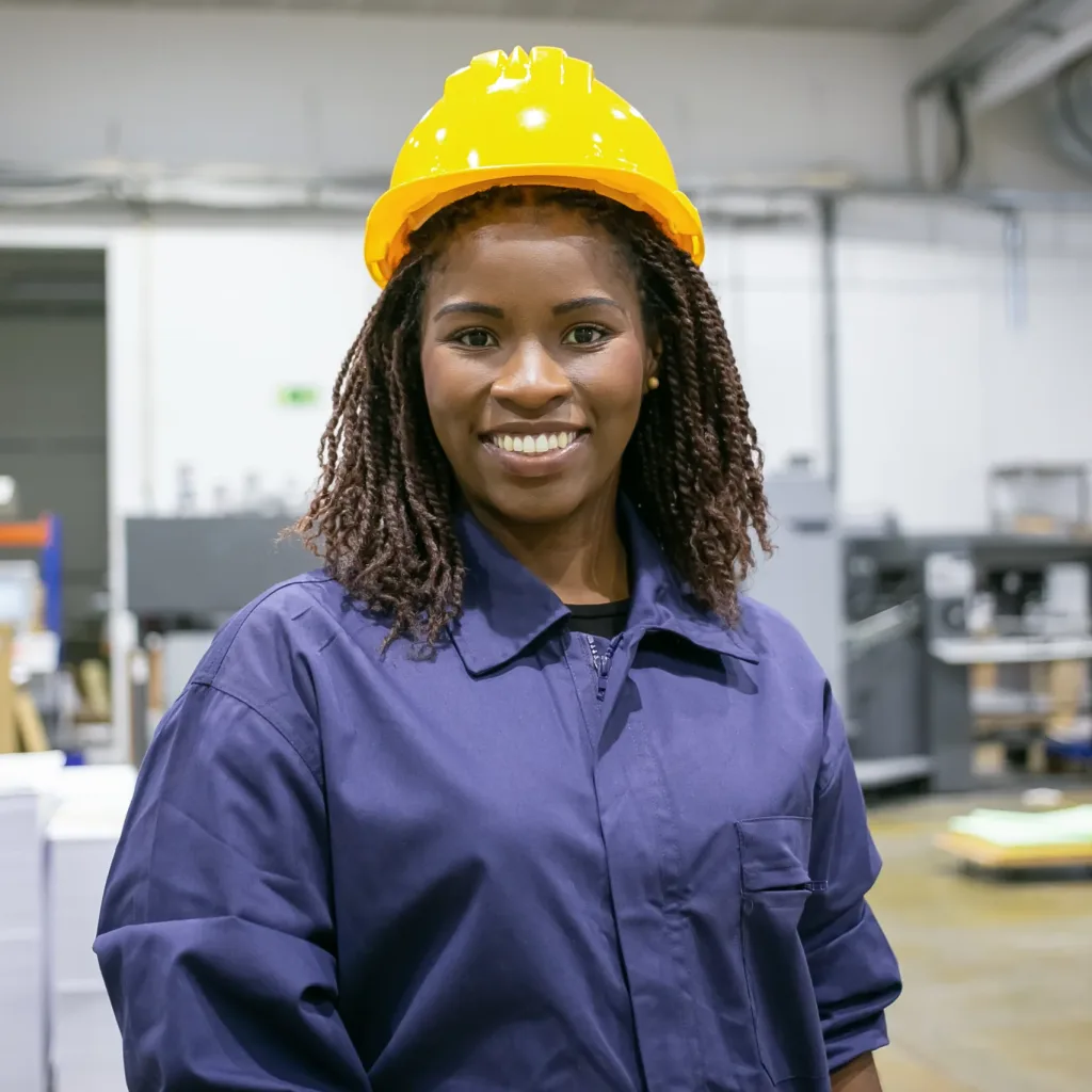 Cheerful African American female factory employee Cheerful African American female factory employee in hardhat and overall standing on plant floor, looking at camera and smiling. Front view, medium shot. Women in industry concept